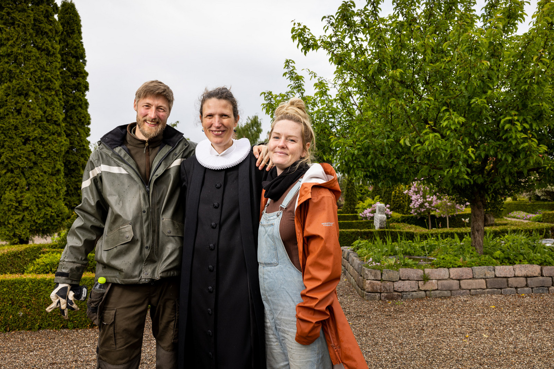 En præst og to medarbejdere smiler sammen i en kirkahave.