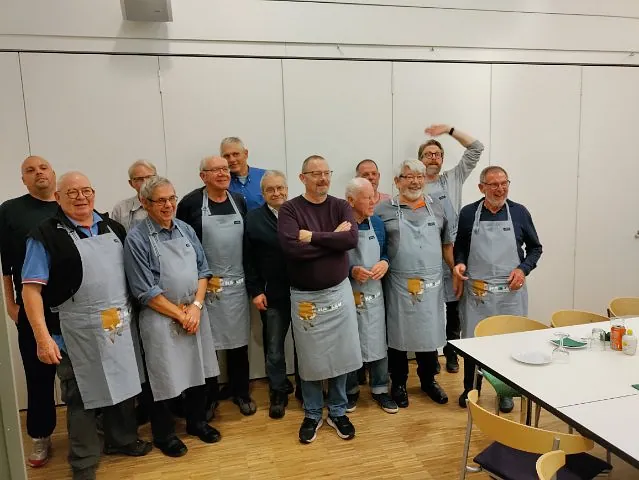 En gruppe mænd i grå "Madklub"-forklæder samlet i sognegården.